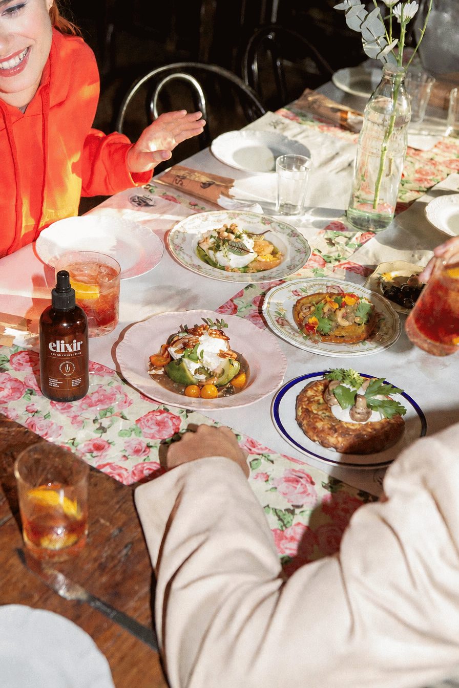 the elixir bottle on a long lunch table with friends, plates and glasses of elixir and tonic