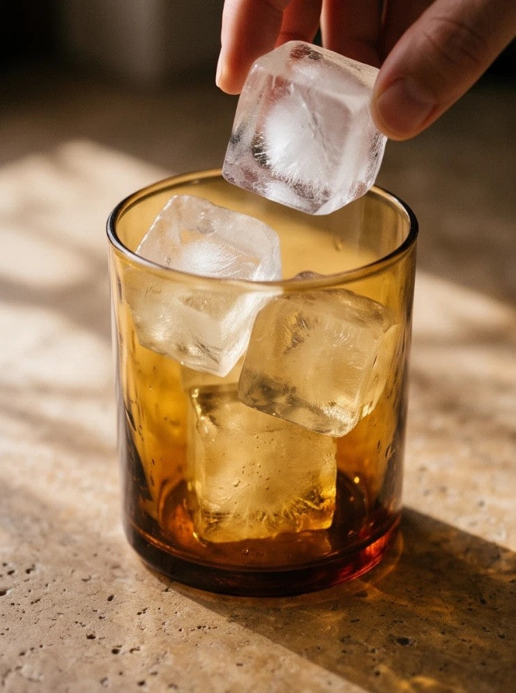 a hand dropping ice into an amber glass